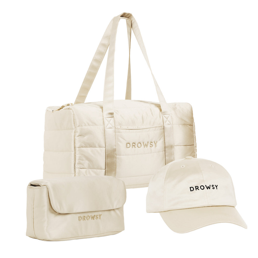 The Drowsy Weekend Collection in Dusty Gold includes a quilted duffle, travel pouch, and baseball cap—all with "DROWSY" embroidery. Perfect cream-colored travel essentials for your next getaway.