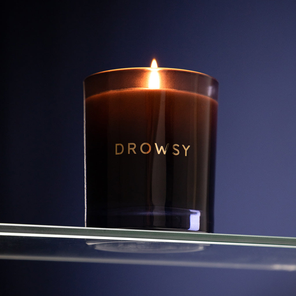 The Sleep Ritual Candle 220g by drowsysleepco rests on a glass shelf against a dark blue background, its dark glass holder inviting relaxation and making it perfect for enhancing your pre-sleep rituals.