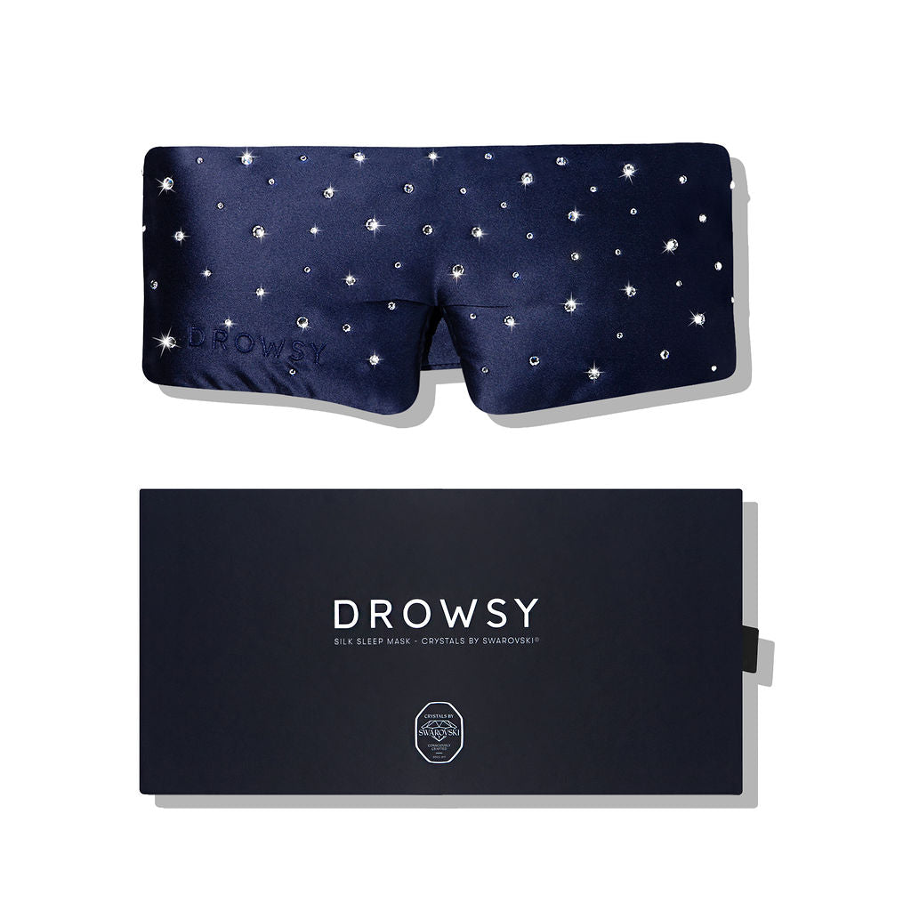 The Silk Sleep Eye Mask Crystals by Swarovski® from us.drowsysleepco is a navy blue silk mask with 100% blackout, adorned with Swarovski crystals, shown atop a matching dark blue box labeled DROWSY Silk Sleep Mask • Crystals by Swarovski.