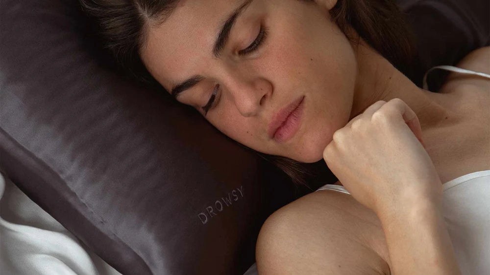 A woman with brown hair sleeps peacefully on her side, resting her head on a dark-colored pillow with the word DROWSY embroidered on it. She wears a white camisole and appears relaxed and comfortable.