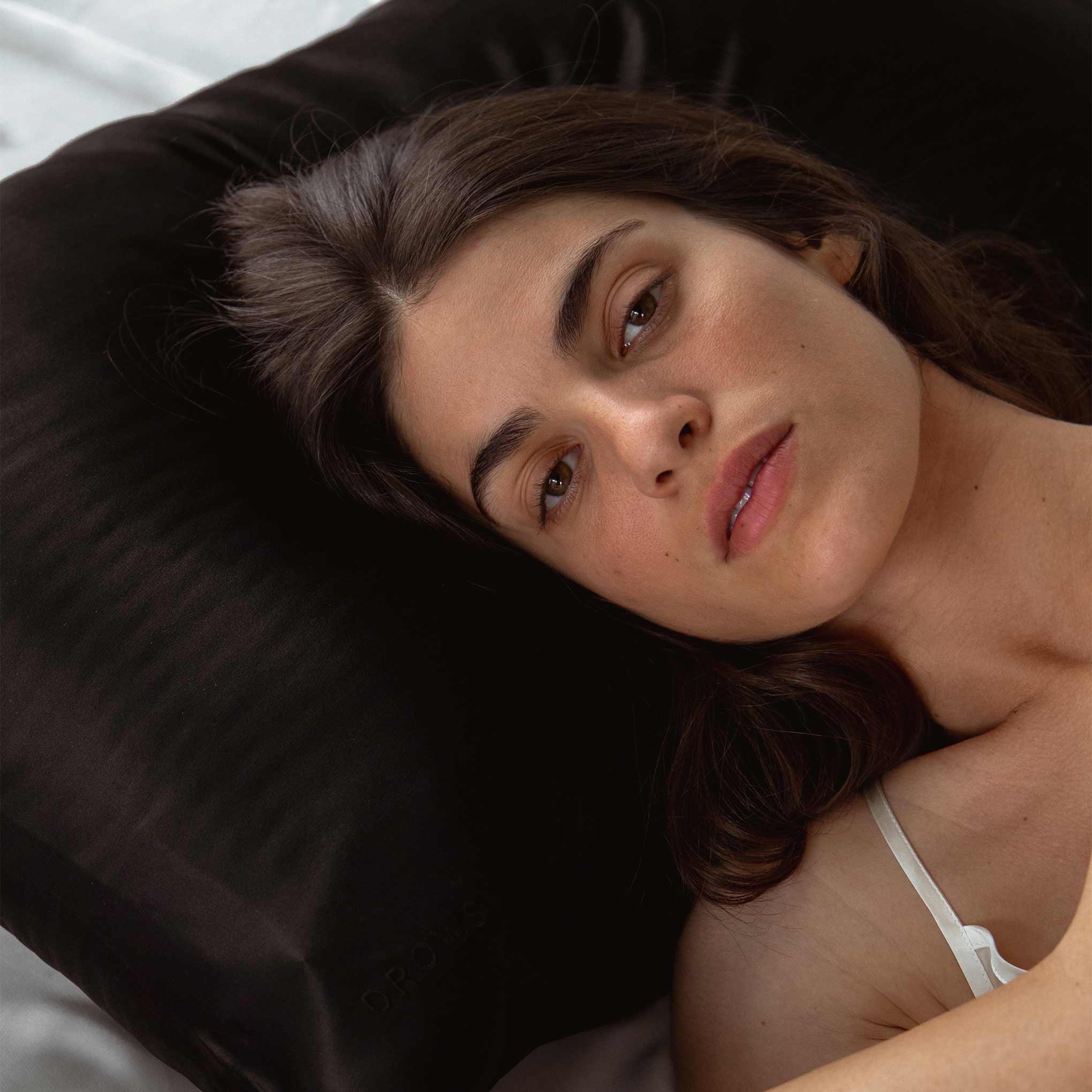 A woman with long dark hair lies on a Black Jade pillow from drowsysleepco’s Drowsy is the New Black Bundle, wearing a white strap top. She looks at the camera in a calm, relaxed setting.