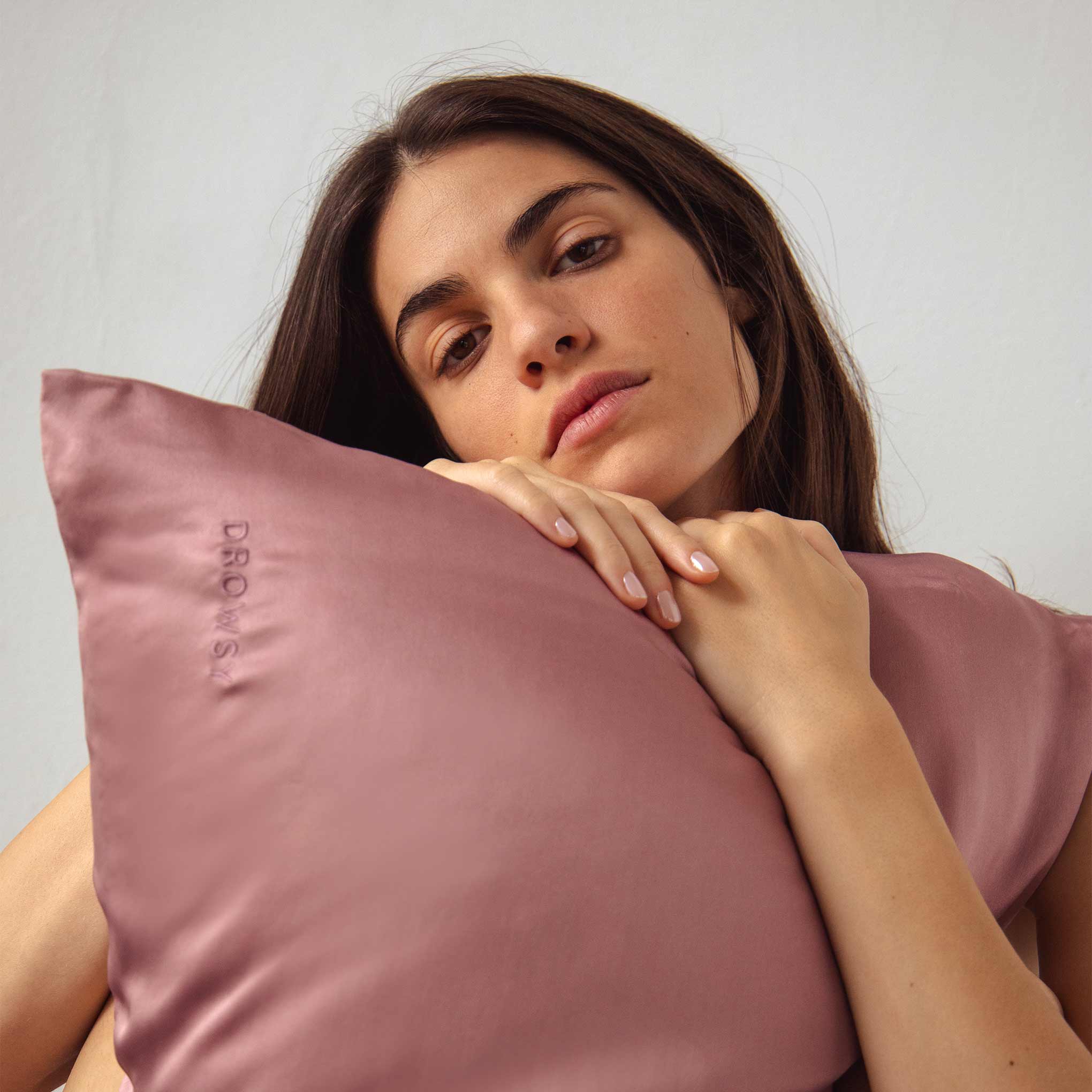 A woman with long brown hair holds and leans her chin on a mauve pillow embroidered with the word DROWSY, looking calmly at the camera against a plain background.