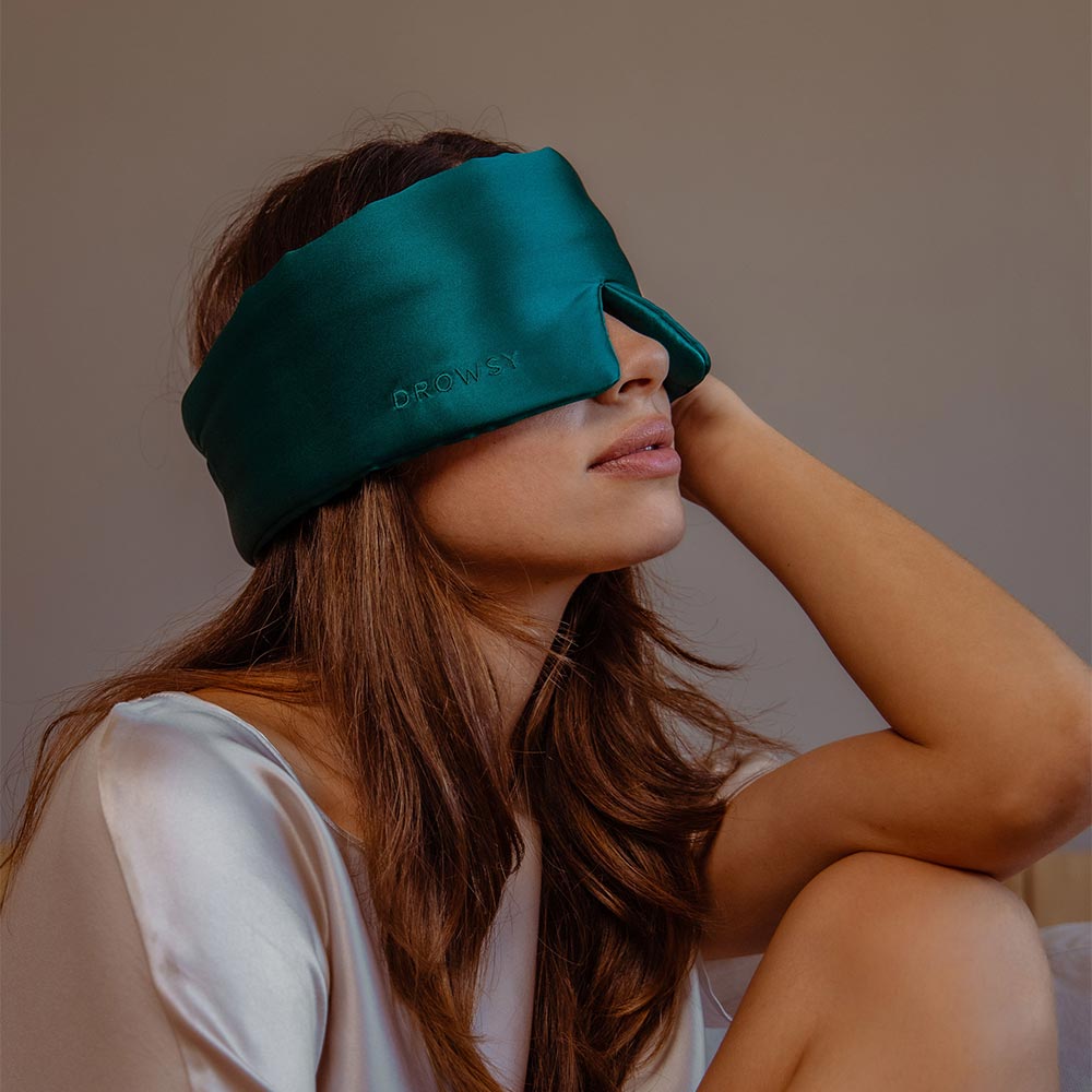 A woman with long brown hair wears a teal satin sleep mask and a silky white top, sitting with one arm resting on her knee against a neutral background.