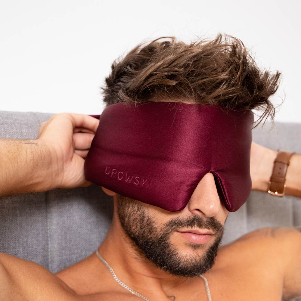 A bearded man adjusts the La Vie En Rouge silk eye mask from us.drowsysleepco while sitting on a bed with a gray upholstered headboard, getting ready for restful beauty sleep.