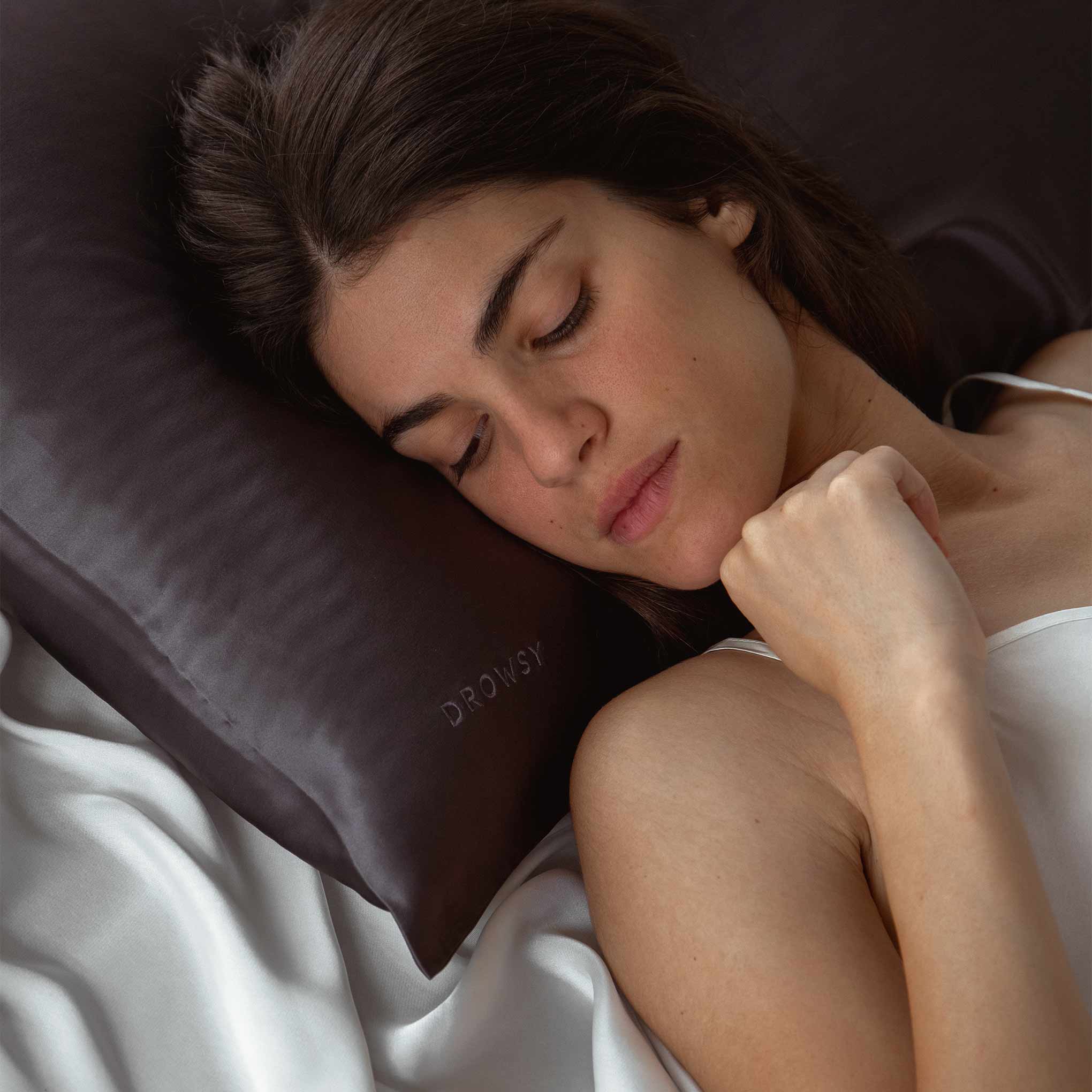 A woman with long dark hair sleeps peacefully on a dark satin pillow that says DROWSY, lying on white satin sheets and wearing a white top.