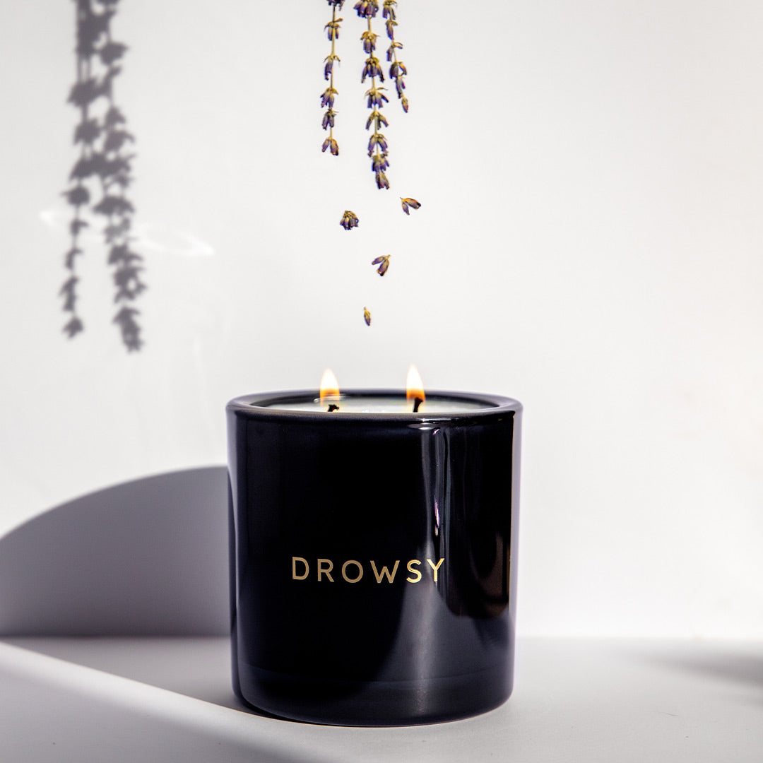 A black candle holder labeled DROWSY with two lit flames sits on a white surface. Sprigs of lavender hang above, casting a delicate shadow on the wall.