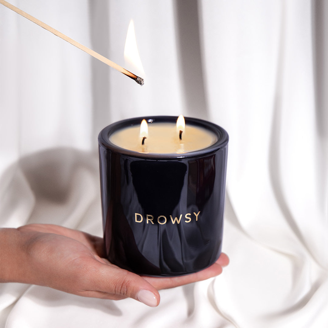 A hand holds a black candle jar labeled DROWSY with three lit wicks, while another hand lights one wick with a match. White fabric drapes in the background.