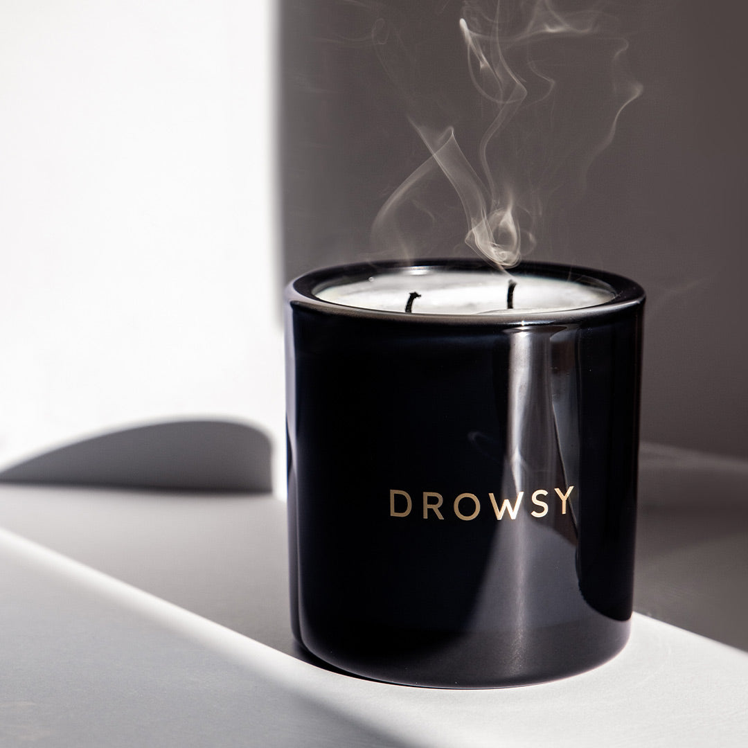 A black candle jar with the word DROWSY in gold letters on the front sits on a light surface. The candle has just been extinguished, with visible wisps of smoke rising from the wicks.