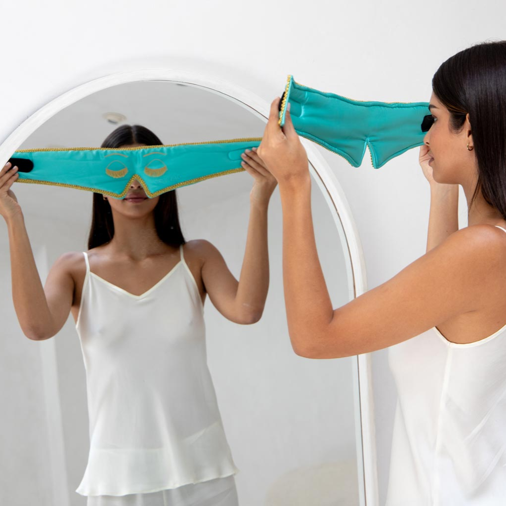 A woman in a white camisole stands before a mirror, holding up a drowsysleepco 2-Pack Sleep Mask from the Breakfast at Drowsy collection, as if she's about to put it on her face.