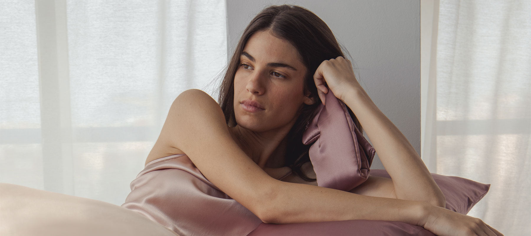 A woman wrapped in a silky light pink sheet reclines on a bed, leaning on a mauve satin pillow and looking thoughtfully to the side. Soft natural light filters through sheer curtains in the background.