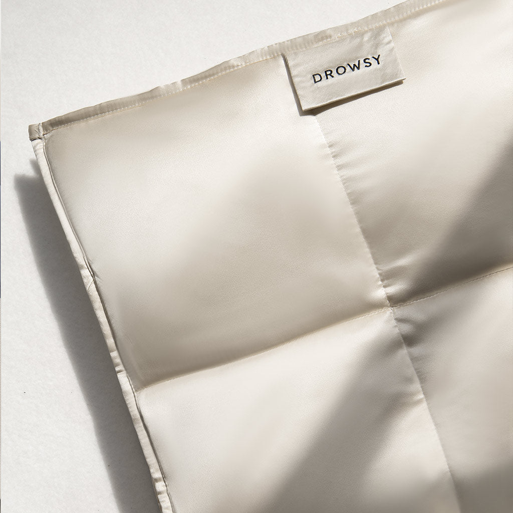 A close-up of the Dusty Gold Silk Weighted Blanket by us.drowsysleepco reveals its light cream, silky quilted fabric with a DROWSY label, large smooth squares, and luxurious shine—promising deep relaxation and an unrivaled sleep experience.