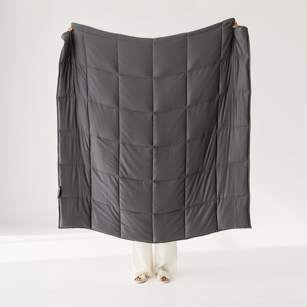 In a white room, a person holds up the us.drowsysleepco Moonlight Shadow Silk Weighted Blanket, covering most of their body and leaving only their feet visible—ideal for deep relaxation and an unmatched sleeping experience.