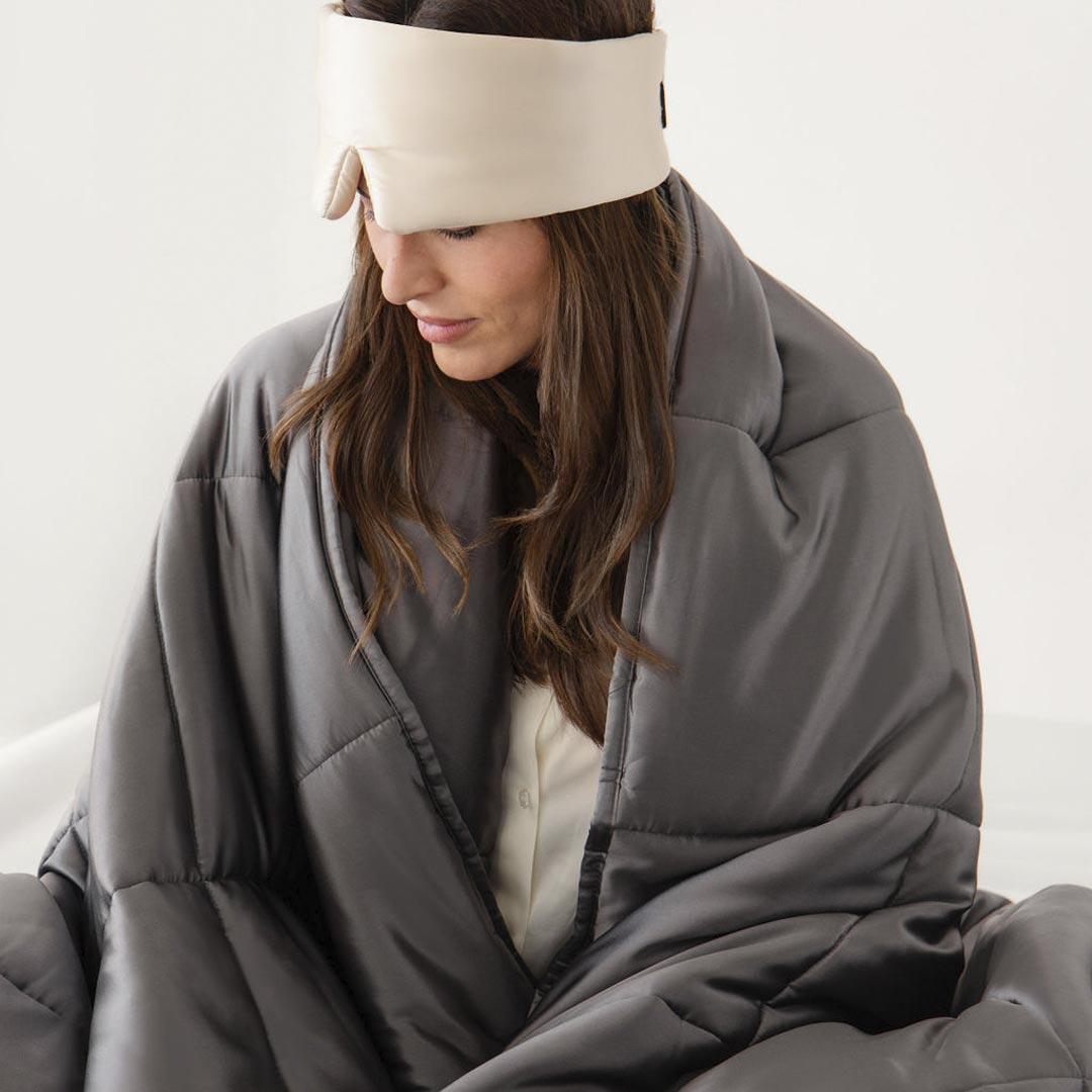 A woman sits wrapped in the us.drowsysleepco Moonlight Shadow Silk Weighted Blanket, wearing a cream eye mask on her forehead and gazing down calmly, embodying deep relaxation.