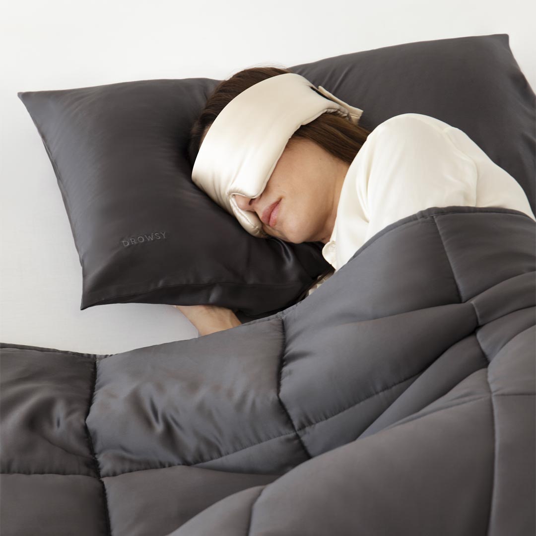A person lies on their side in bed, resting deeply with the us.drowsysleepco Moonlight Shadow Silk Weighted Blanket and a matching gray pillow while wearing a white sleep mask.