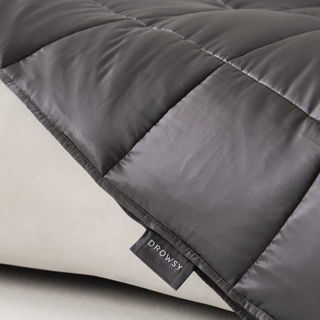 A close-up of the Moonlight Shadow Silk Weighted Blanket by us.drowsysleepco, quilted in dark gray silk with a Drowsy tag, draped over a light surface—designed for deep relaxation and an exceptional sleep experience.