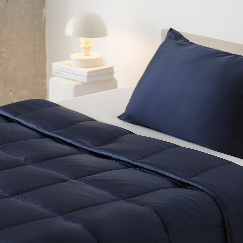 A neatly made bed features navy pillows, a matching comforter, and the us.drowsysleepco Midnight Blue Silk Weighted Blanket for deep relaxation, set beside a nightstand with stacked books and a white round lamp against a light wall.