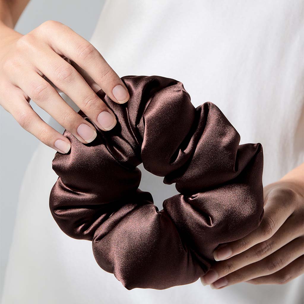 A person in a white top holds the us.drowsysleepco Cocoa Glow - Padded Scrunchie, an ideal hair sleep accessory, with both hands against a neutral background.