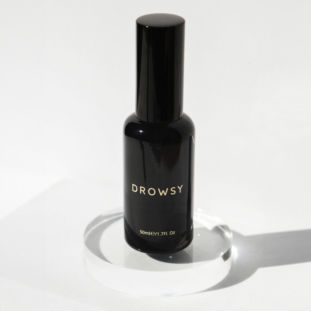 A black bottle labeled DROWSY stands upright on a clear, round platform against a white background. The bottle has a shiny finish and minimalist design, with text showing 50ml/1.7fl.oz beneath the brand name.
