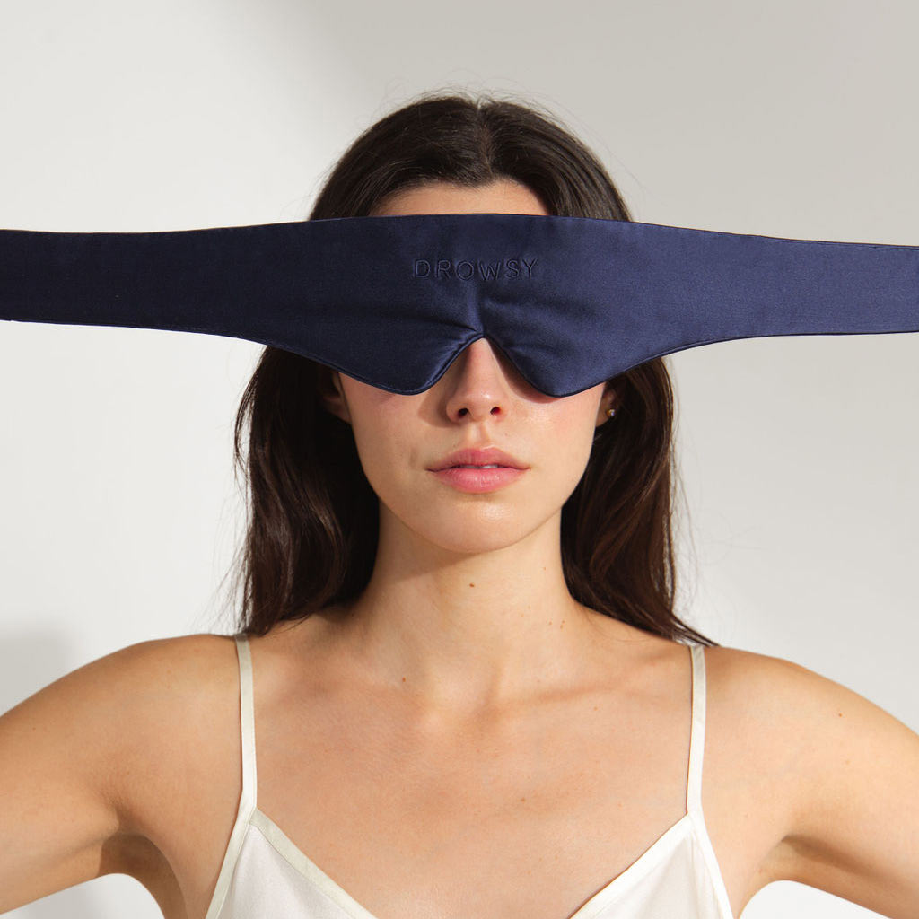 A woman wearing a white camisole poses against a plain background with the us.drowsysleepco Featherweight Silk Eye Mask in Midnight Blue, a blackout design that covers her eyes and extends across her temples.