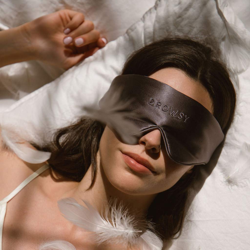 A woman lies on a bed with white sheets, wearing a large silk sleep mask labeled Drowsy. Soft feathers are scattered around her, and sunlight gently illuminates the scene.