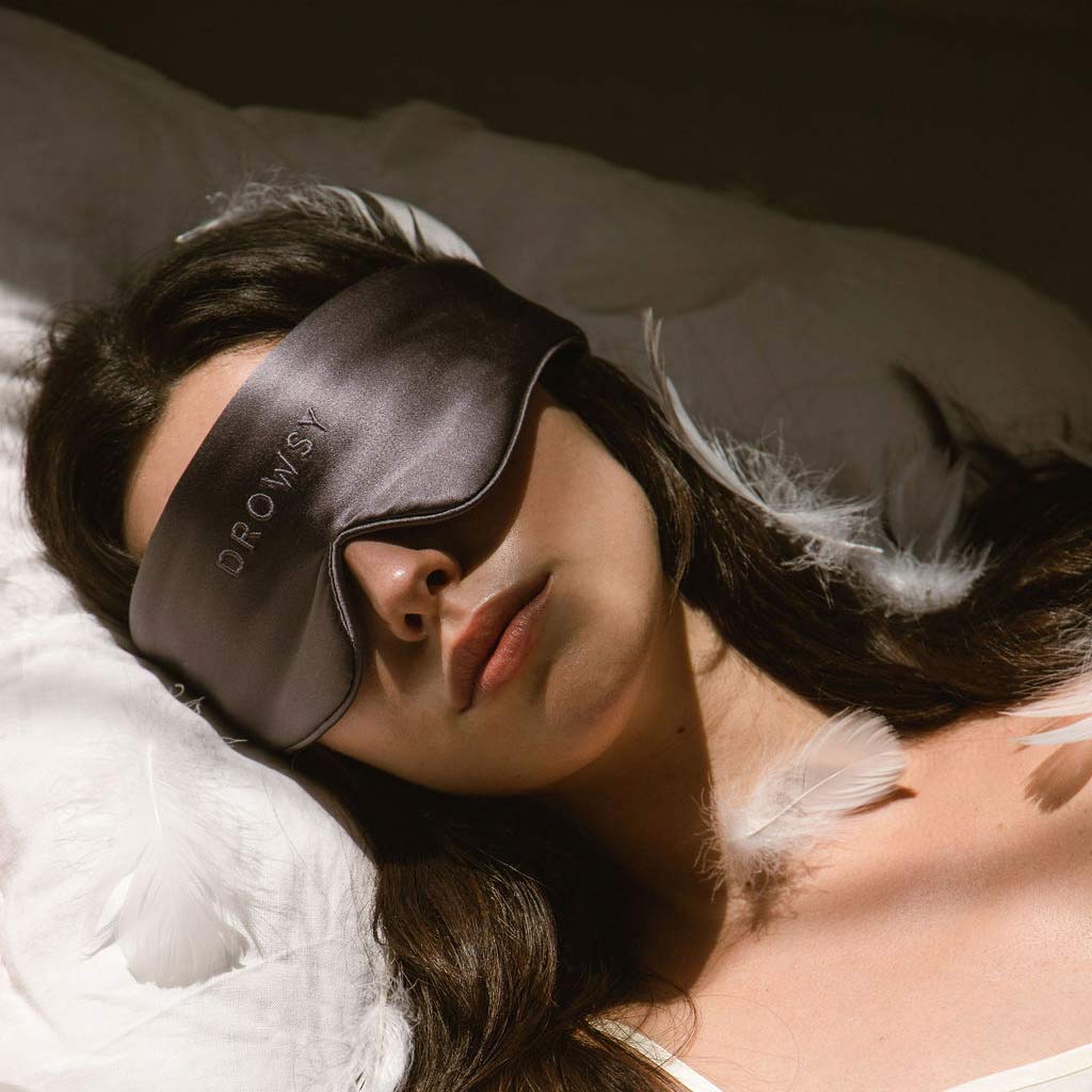 A woman with long dark hair lies on a white pillow, wearing a gray sleep mask that says DROWSY. Soft white feathers are scattered around her as she rests peacefully in natural light.