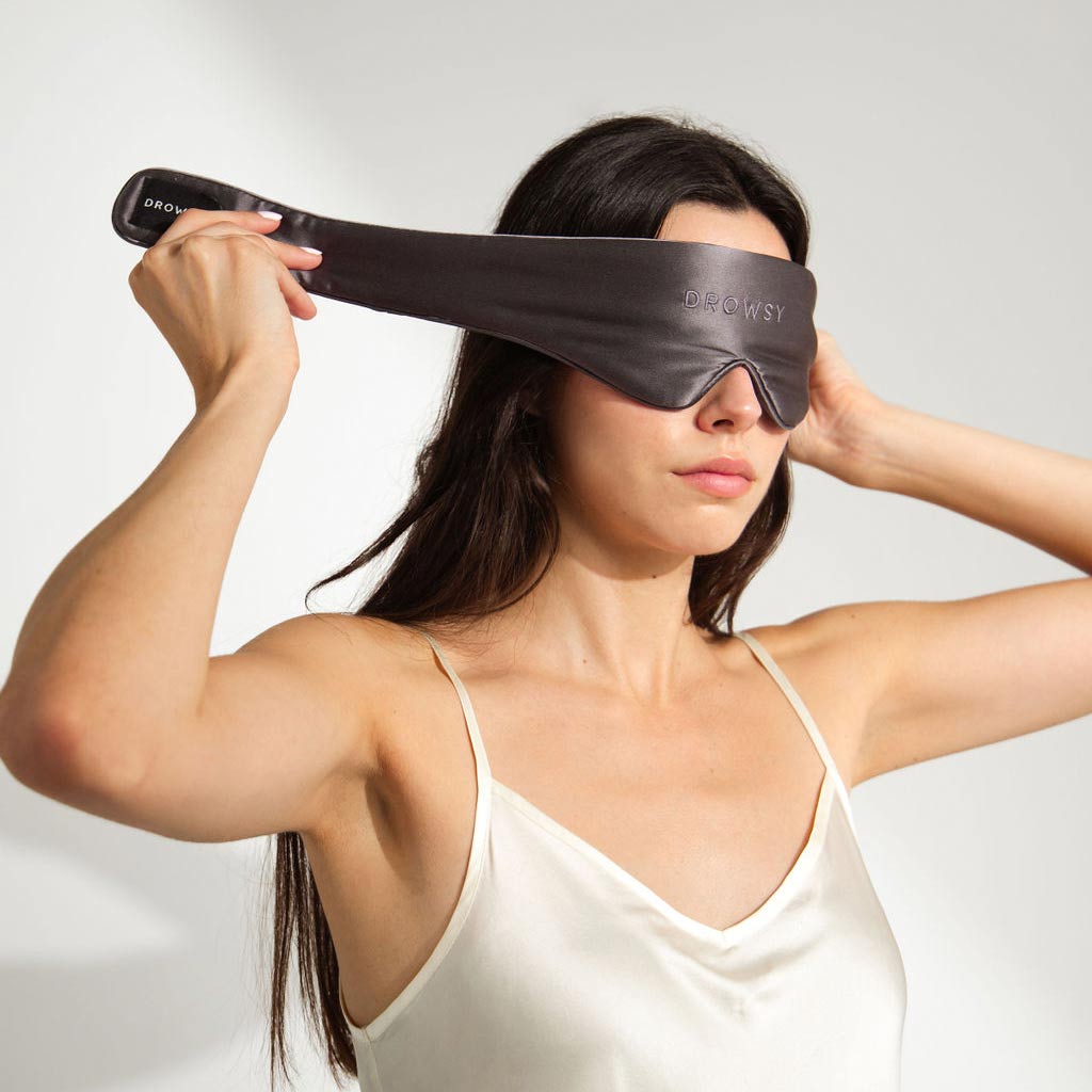 A woman in a white satin camisole puts on a black sleep mask labeled DROWSY. She is standing against a plain light background, and her long dark hair is loose.