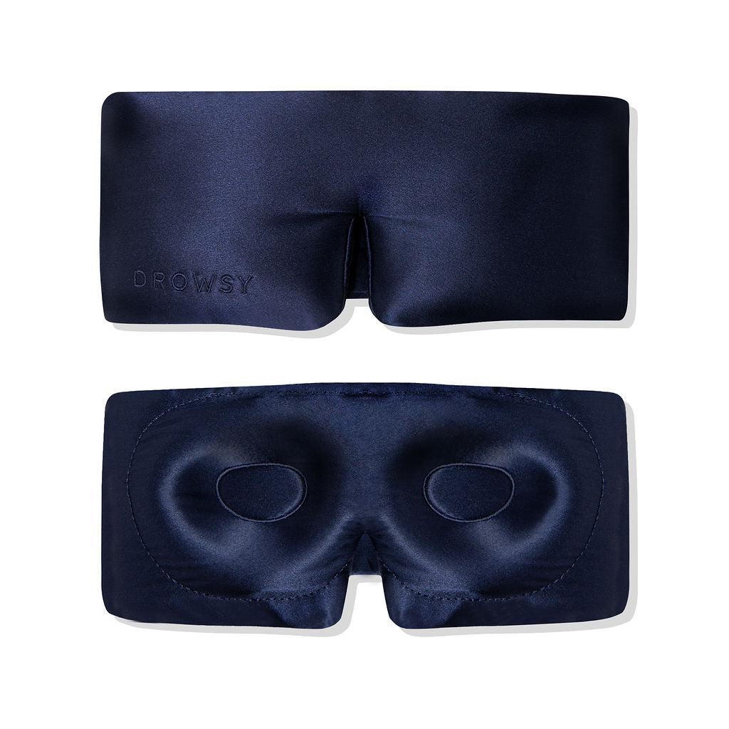 Two dark blue silk sleep masks are shown; one displays the outer side with DROWSY embossed, and the other shows the inner side with padded eye contours for comfort. Both are set against a white background.