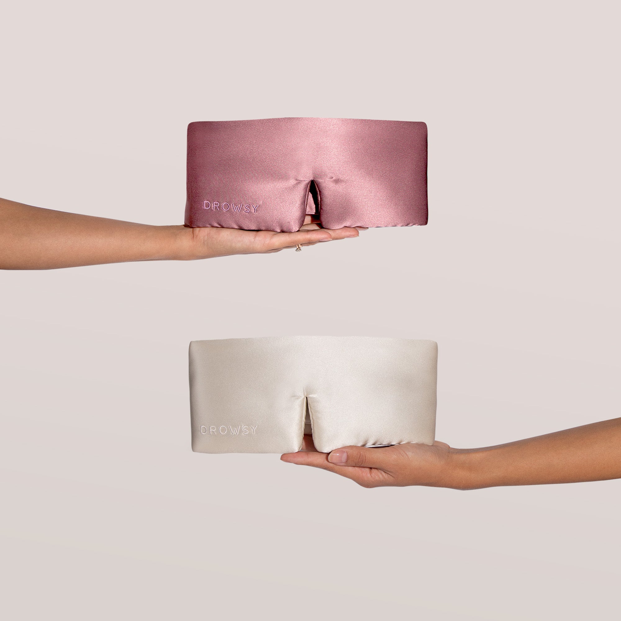 Two hands hold out silk sleep masks in pink (top) and cream (bottom), each embroidered with the word DROWSY on the side, against a light, plain background.