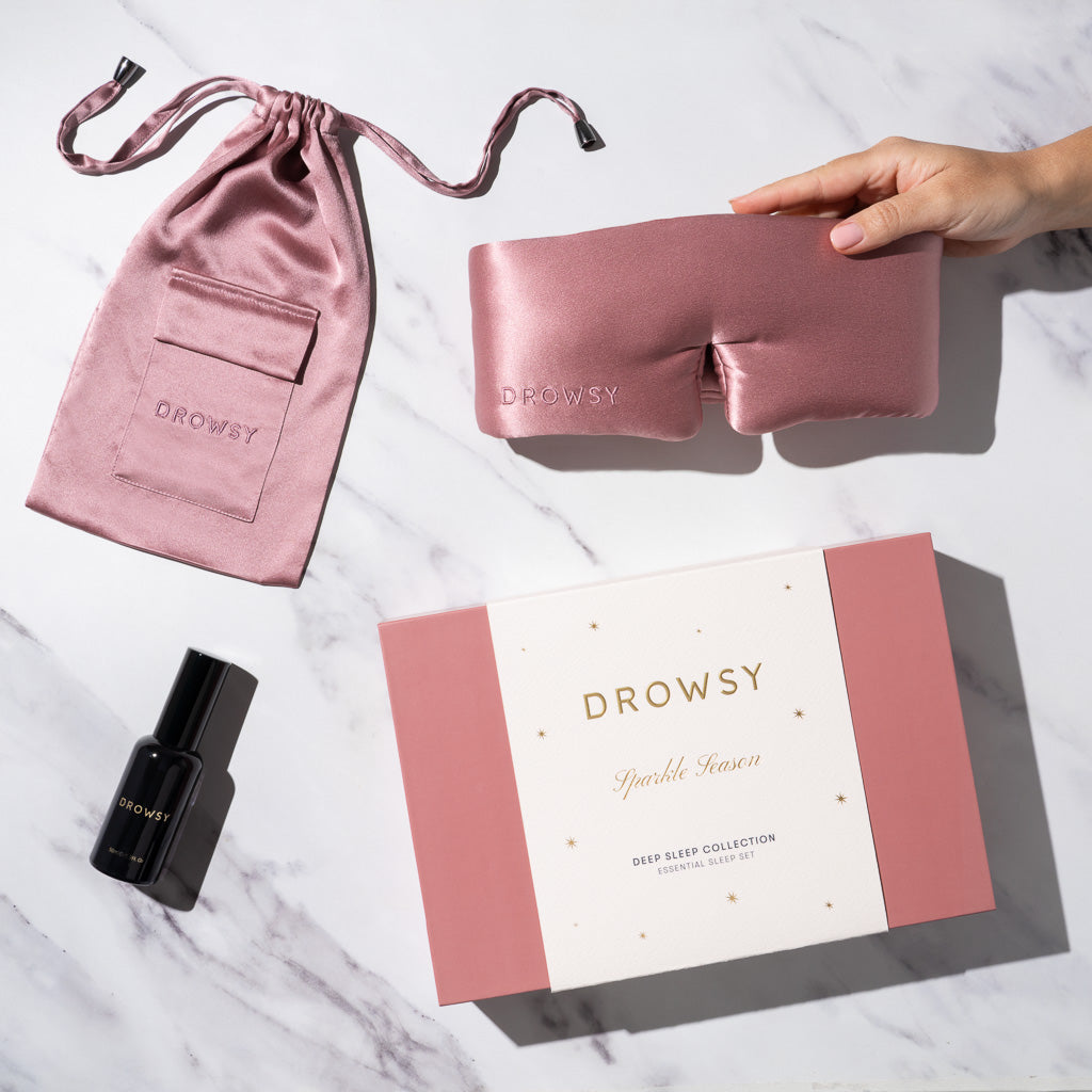 A hand holds the Damask Rose - Deep Sleep Collection sleep mask by us.drowsysleepco, paired with a matching satin pouch, deep sleep pillow spray in a sleek black bottle, and a pink and white gift box on white marble.