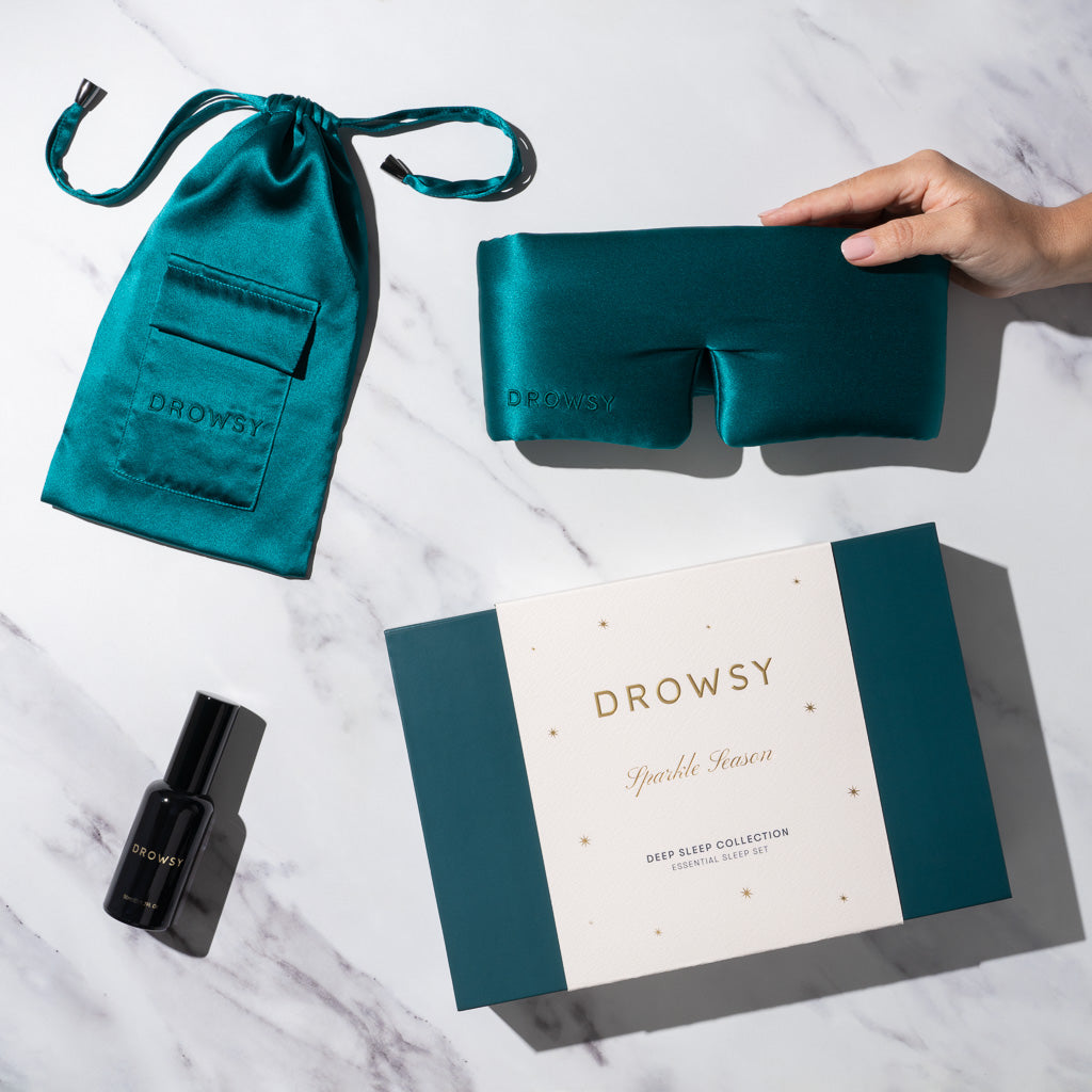A hand holds the Green Sapphire - Deep Sleep Collection sleep mask by us.drowsysleepco, with its silk mask, matching pouch, and black pillow spray arranged on marble beside the branded box—dreamy sleep essentials at their best.