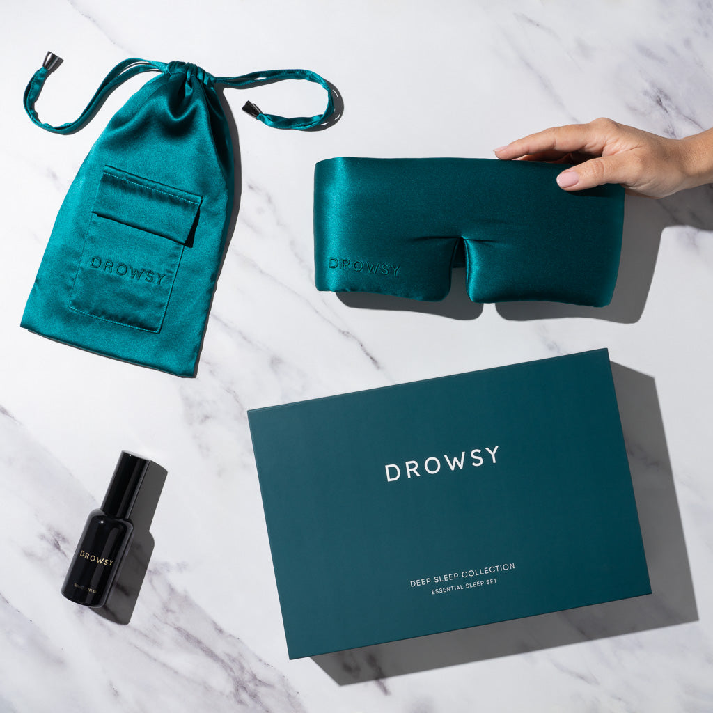A hand holds us.drowsysleepco's Green Sapphire - Deep Sleep Collection: a luxury teal silk sleep mask, matching pouch, deep sleep pillow spray in a black bottle, and teal gift box, all displayed on white marble.