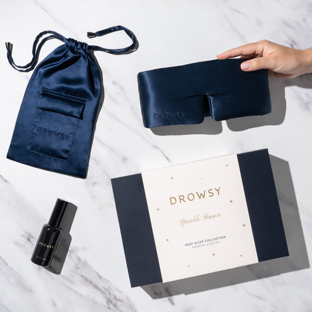 A hand holds the us.drowsysleepco Midnight Blue - Deep Sleep Collection: a luxury silk sleep mask, matching blue silk carry pouch, deep sleep pillow spray in a black bottle, and gift box on a white marble surface.
