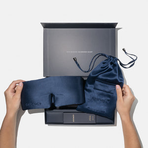 A person holds the us.drowsysleepco Midnight Blue - Deep Sleep Collection silk sleep mask and blue pouch above an open gift box reading "You deserve glorious sleep," with a deep sleep pillow spray inside.