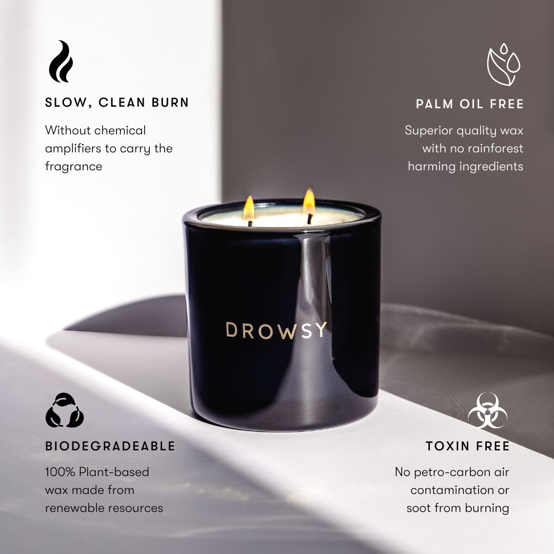 A black candle labeled Drowsy with two flames sits on a white surface. Text around it highlights features: slow, clean burn; palm oil free; biodegradable; and toxin free. Minimalist design with soft shadows.