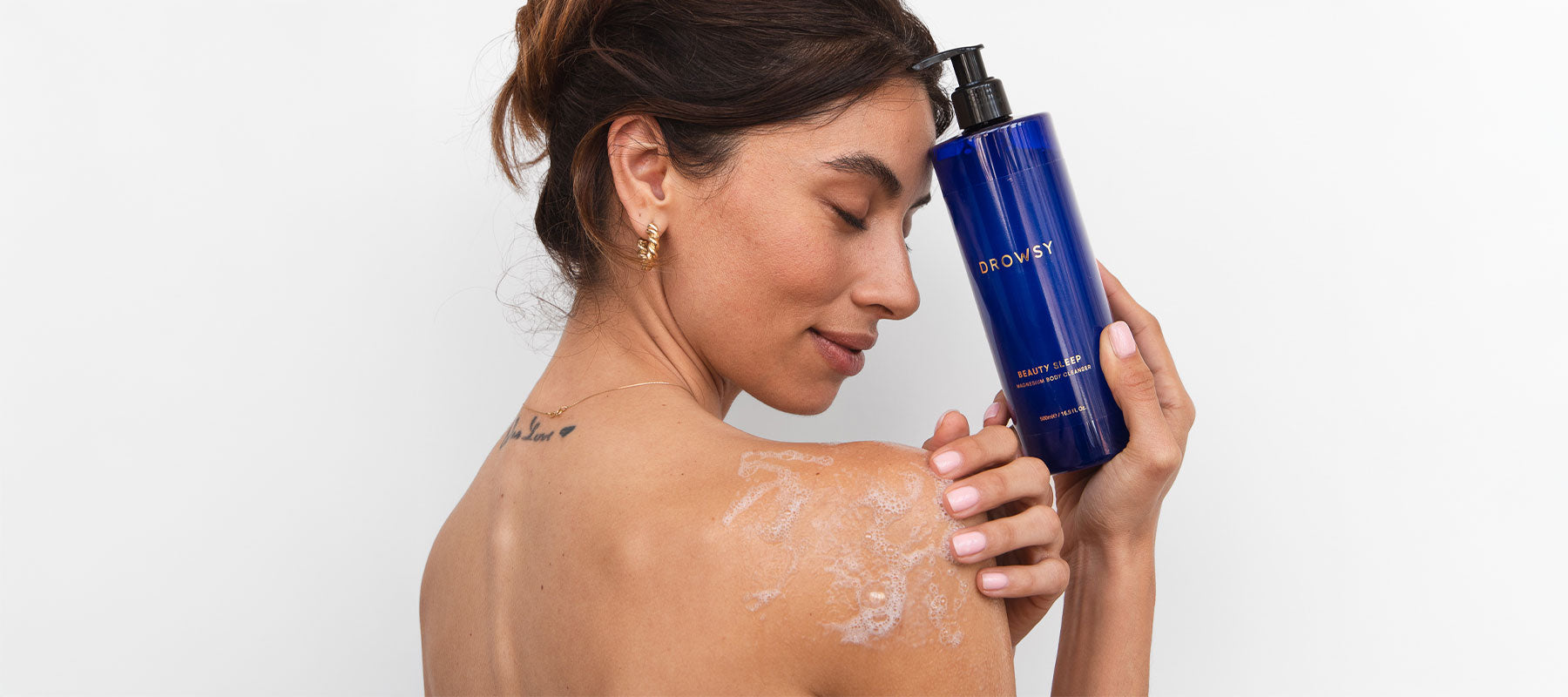 A woman with wavy brown hair applies a foamy lotion to her bare shoulder while holding a blue pump bottle labeled BODY close to her face. She wears gold hoop earrings and stands against a white background.