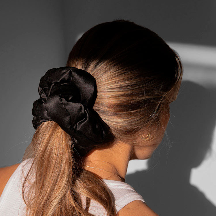 A woman with long, light brown hair wears a large black satin scrunchie in a low ponytail. She is dressed in a white tank top and is facing away from the camera, standing in soft, natural light.