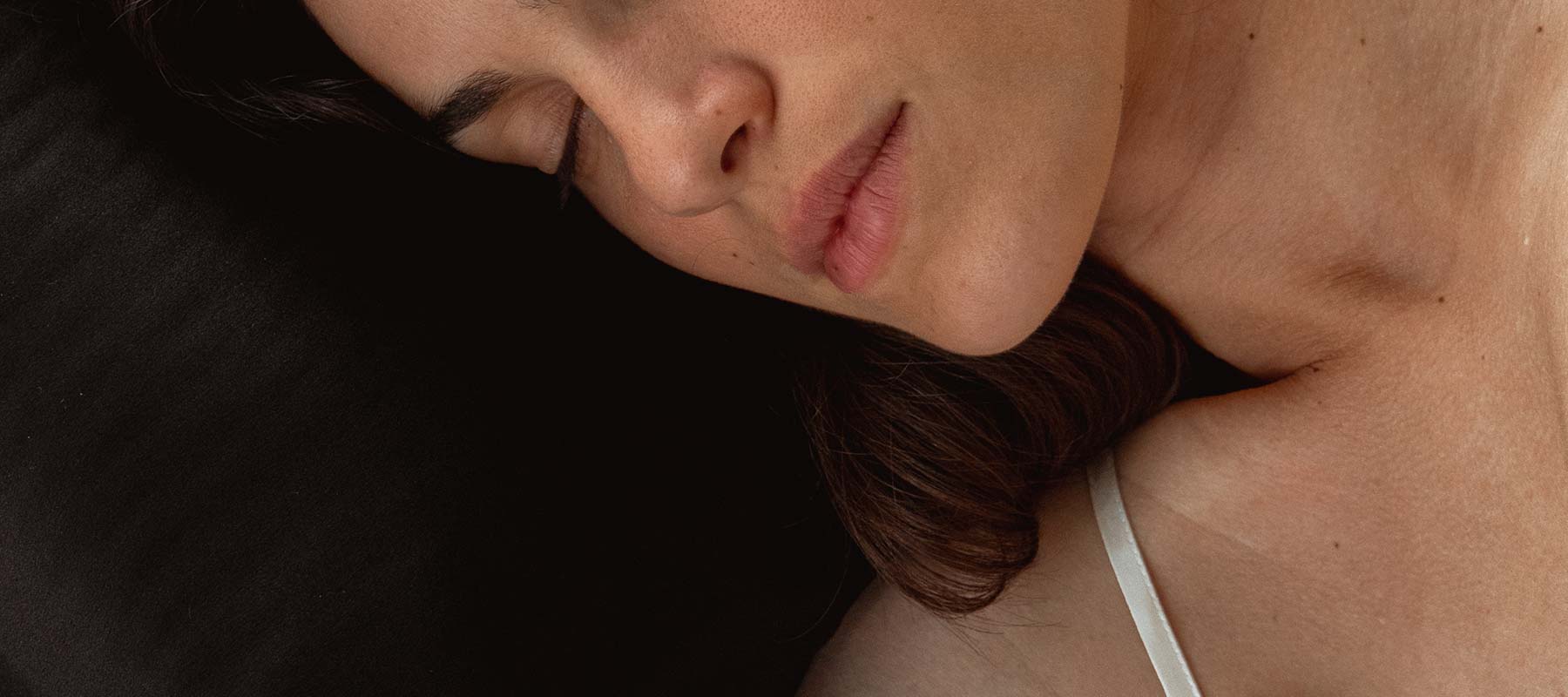 Close-up of a woman with brown hair lying on her side, eyes closed, resting on a black surface while wearing a white strap top.
