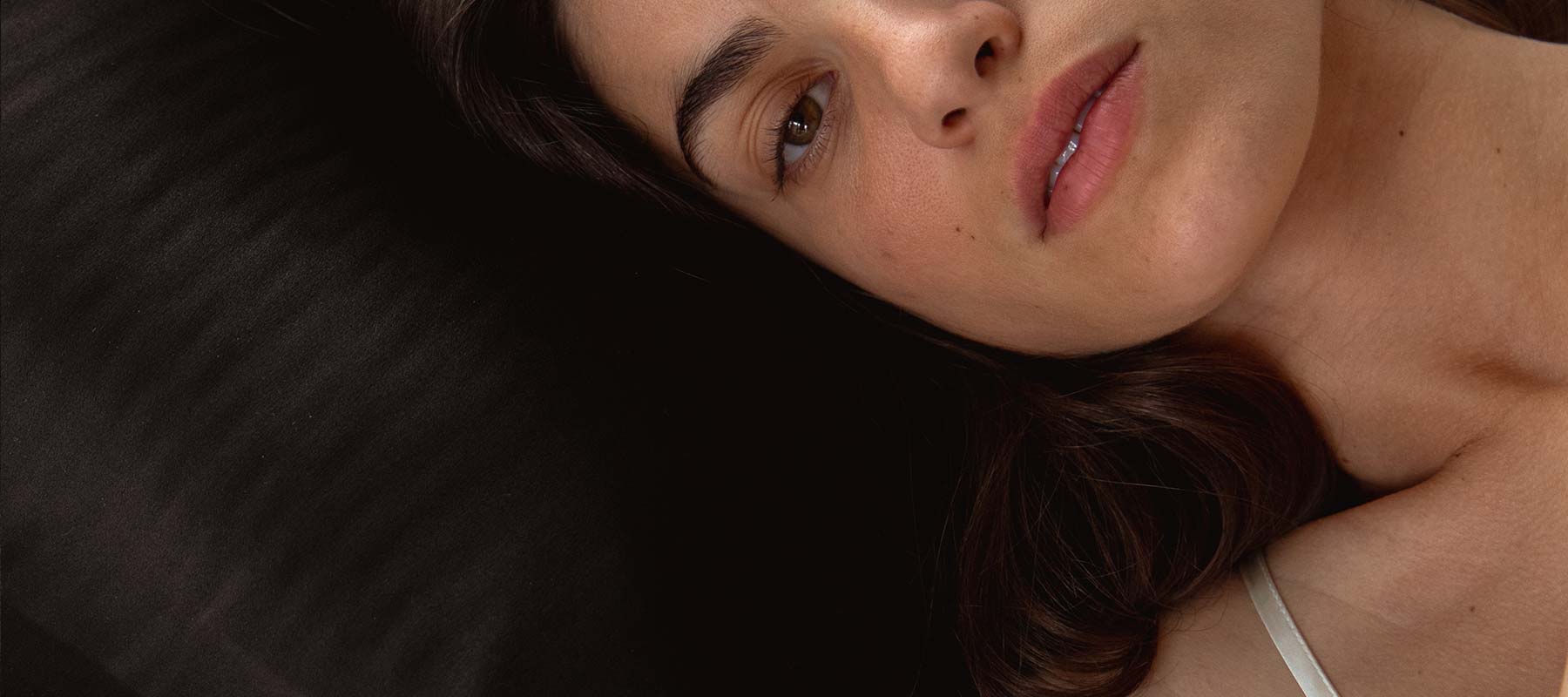A woman with long brown hair lies on her side on a black pillow, looking thoughtfully at the camera. She wears a white top, and her face is partially visible in the close-up image.