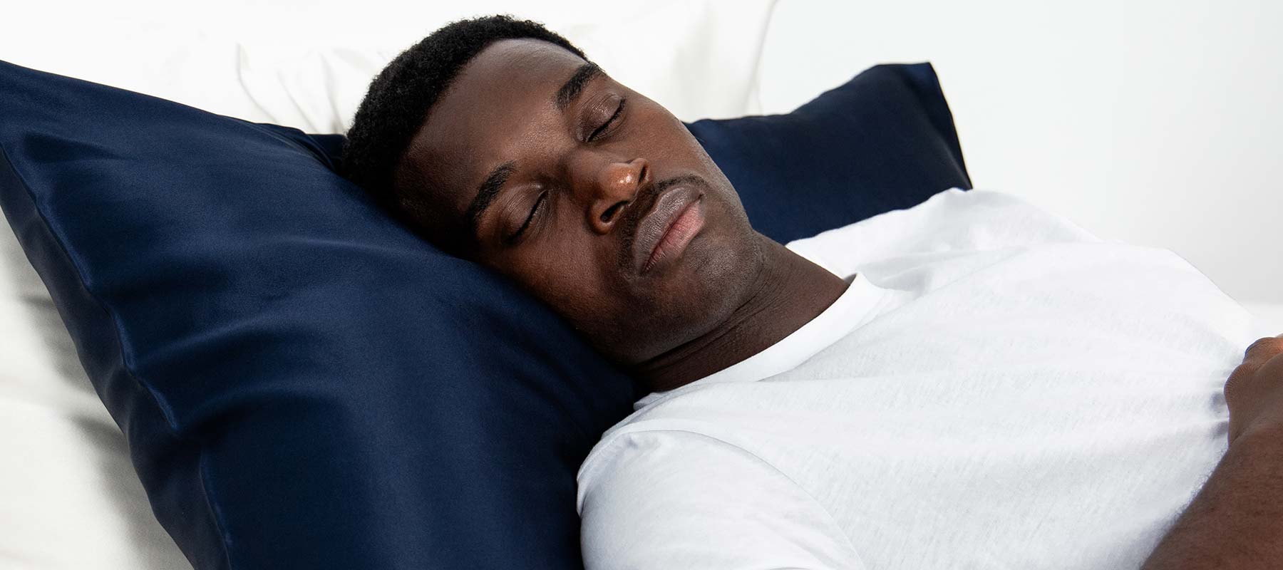 A man wearing a white t-shirt is lying on his back, sleeping peacefully on a bed with a dark blue pillow and white sheets.