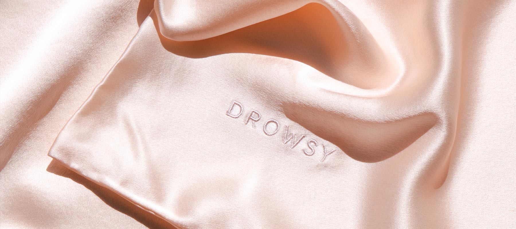 A light peach-colored silk fabric with the word DROWSY embroidered near the edge, softly folded and reflecting gentle light.