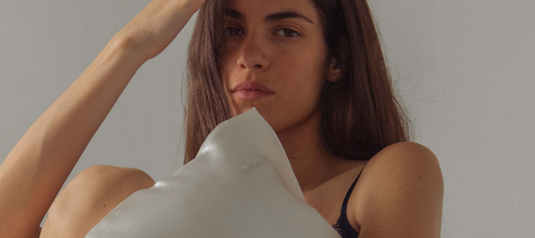 A woman with long brown hair holds a white pillow close to her chest. She looks at the camera with a neutral expression, one hand touching her head. The background is plain and softly lit.
