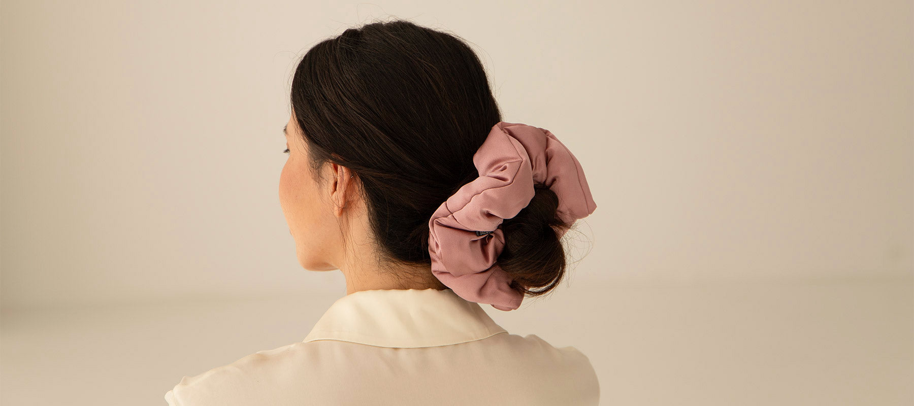 A woman with dark hair styled in a low bun, secured with a large dusty-pink scrunchie, is seen from behind against a plain, light-colored background. She is wearing a cream-colored collared shirt.