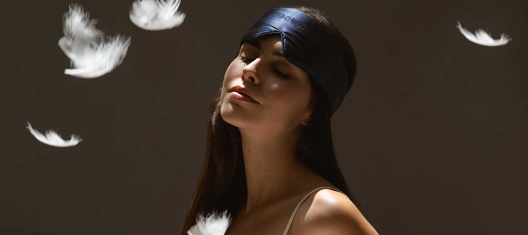 A woman wearing a black sleep mask tilts her head back with a relaxed expression as white feathers float around her against a dark background.