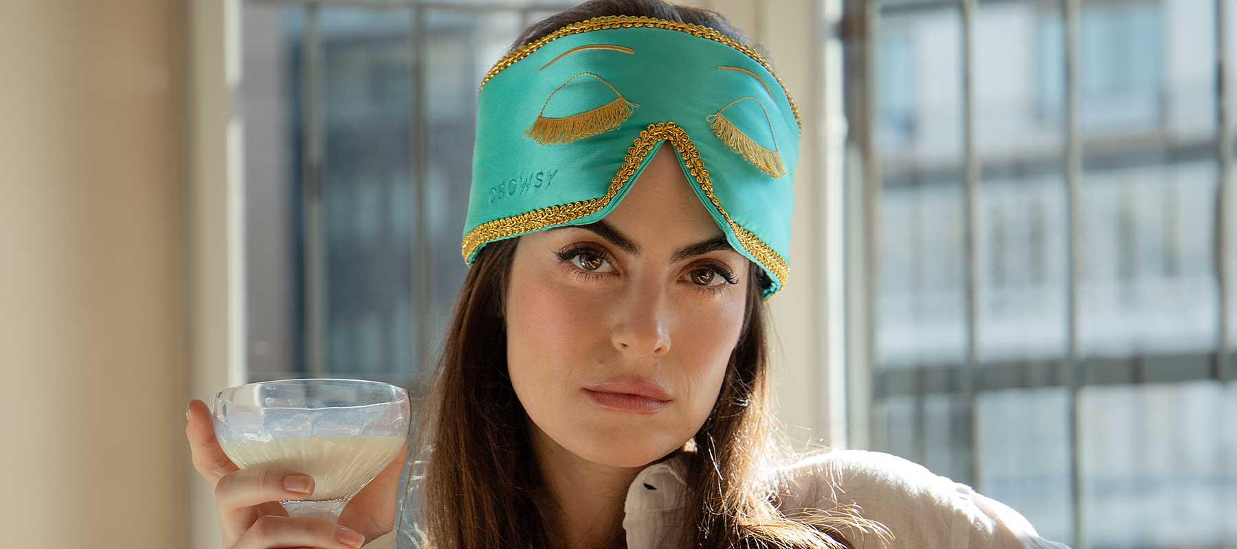 A woman with long brown hair wears a turquoise sleep mask with gold trim pushed up on her forehead, holding a glass of a creamy beverage, with a neutral expression, indoors by a window with bars.