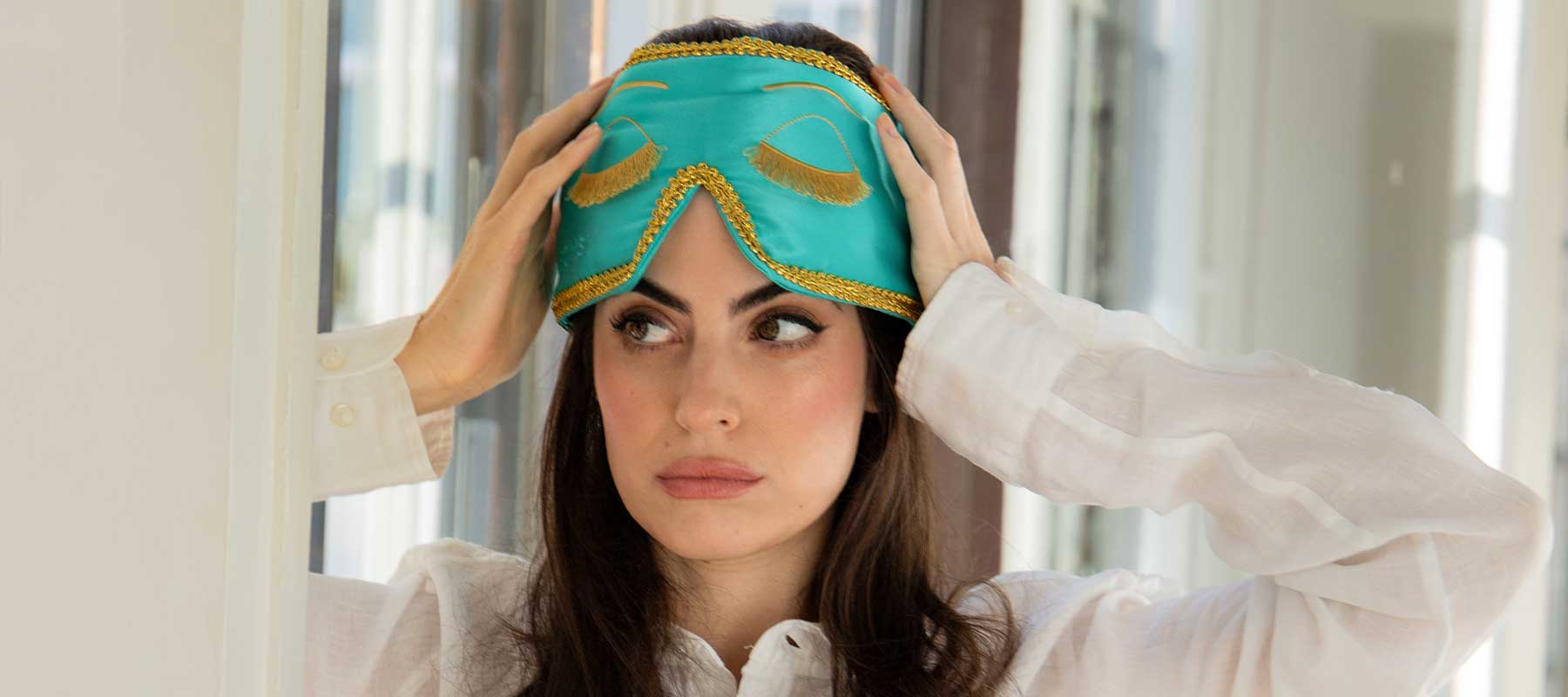 A woman with long dark hair in a white shirt holds a teal and gold sleep mask on her head, gazing thoughtfully to the side in a brightly lit room.