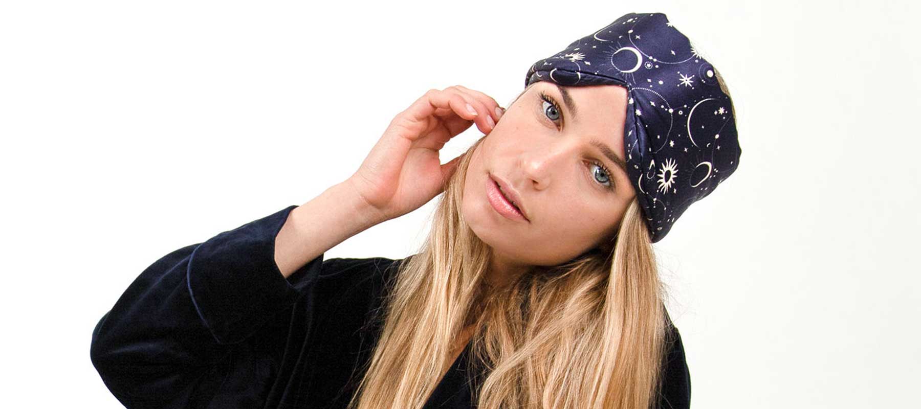 A woman with long blonde hair wears a dark blue headscarf decorated with white celestial designs, including moons and stars. She is dressed in a dark top and tilts her head slightly, touching her ear.