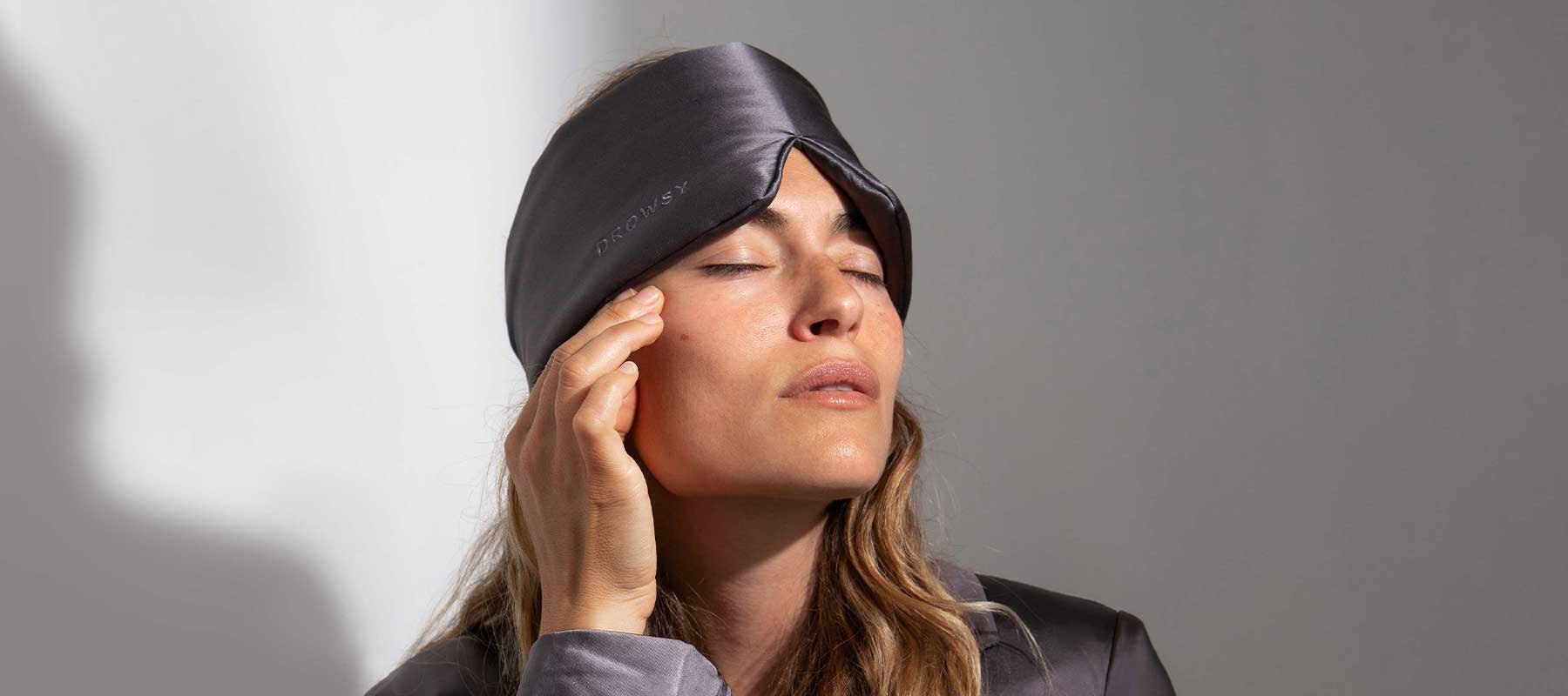 A woman wearing a gray satin sleep mask tilts her head slightly and touches her temple with her fingers, eyes closed, appearing calm and relaxed.