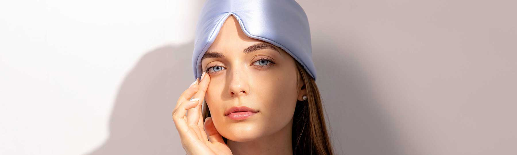 A woman with long brown hair wearing a shiny light blue sleep mask lifted on her forehead gently touches her face with her hand, looking directly at the camera against a neutral background.