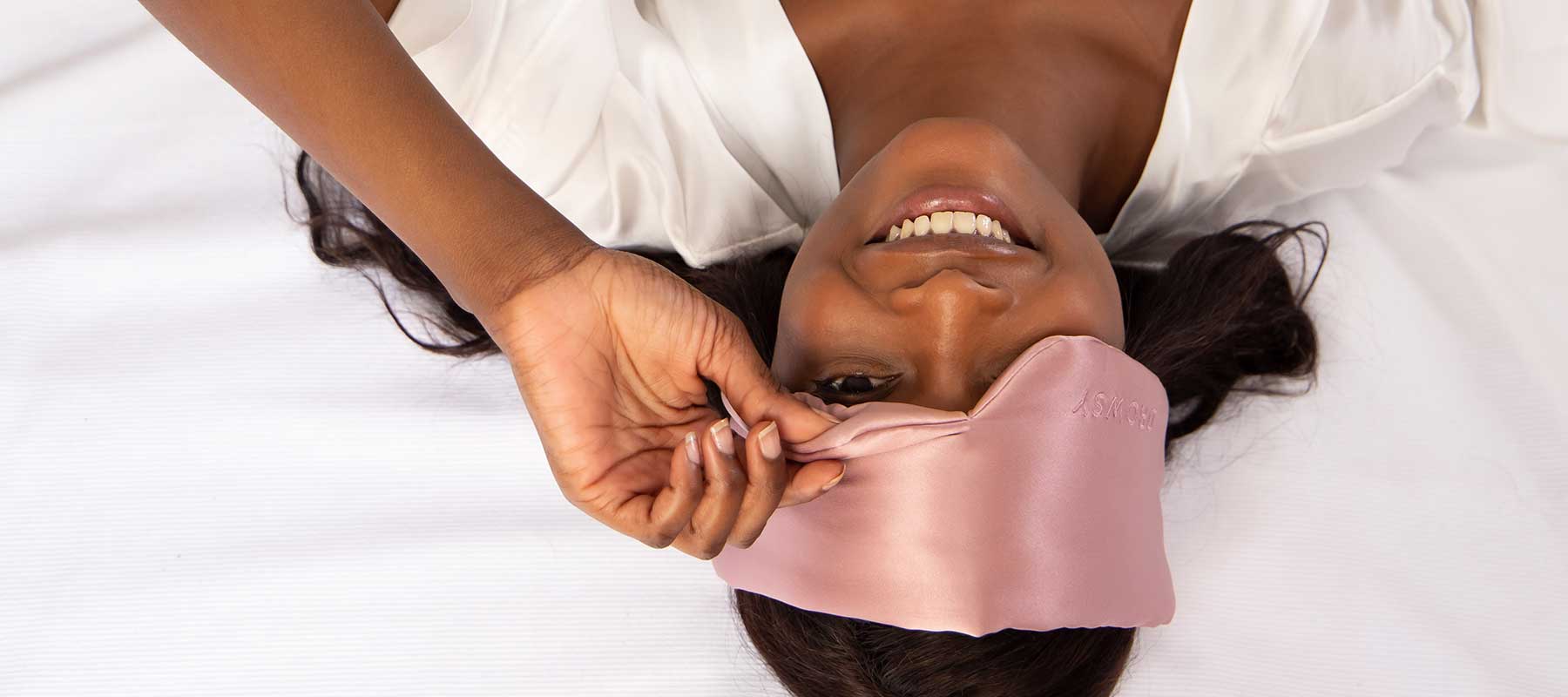 A woman lies on her back on a bed, smiling, while lifting a pink satin sleep mask from her eyes. She is wearing a white robe and appears to be relaxed and happy.