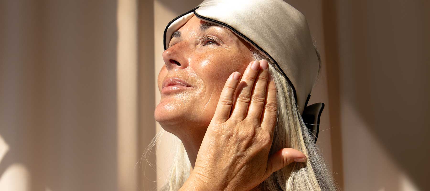 An older woman wearing a satin sleep mask tilted on her head smiles softly while touching her cheek, basking in warm natural sunlight.